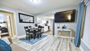Una sala moderna con comedor, un televisor de pantalla plana montado en la pared y una cocina al fondo.