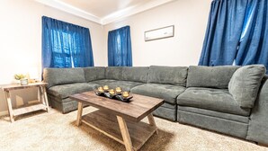 A living room with a grey sectional sofa, a wooden coffee table with candles, a small side table, and blue curtains. - Comfortable 3-Bedroom Home in a Great Central Location (Lincoln Park)