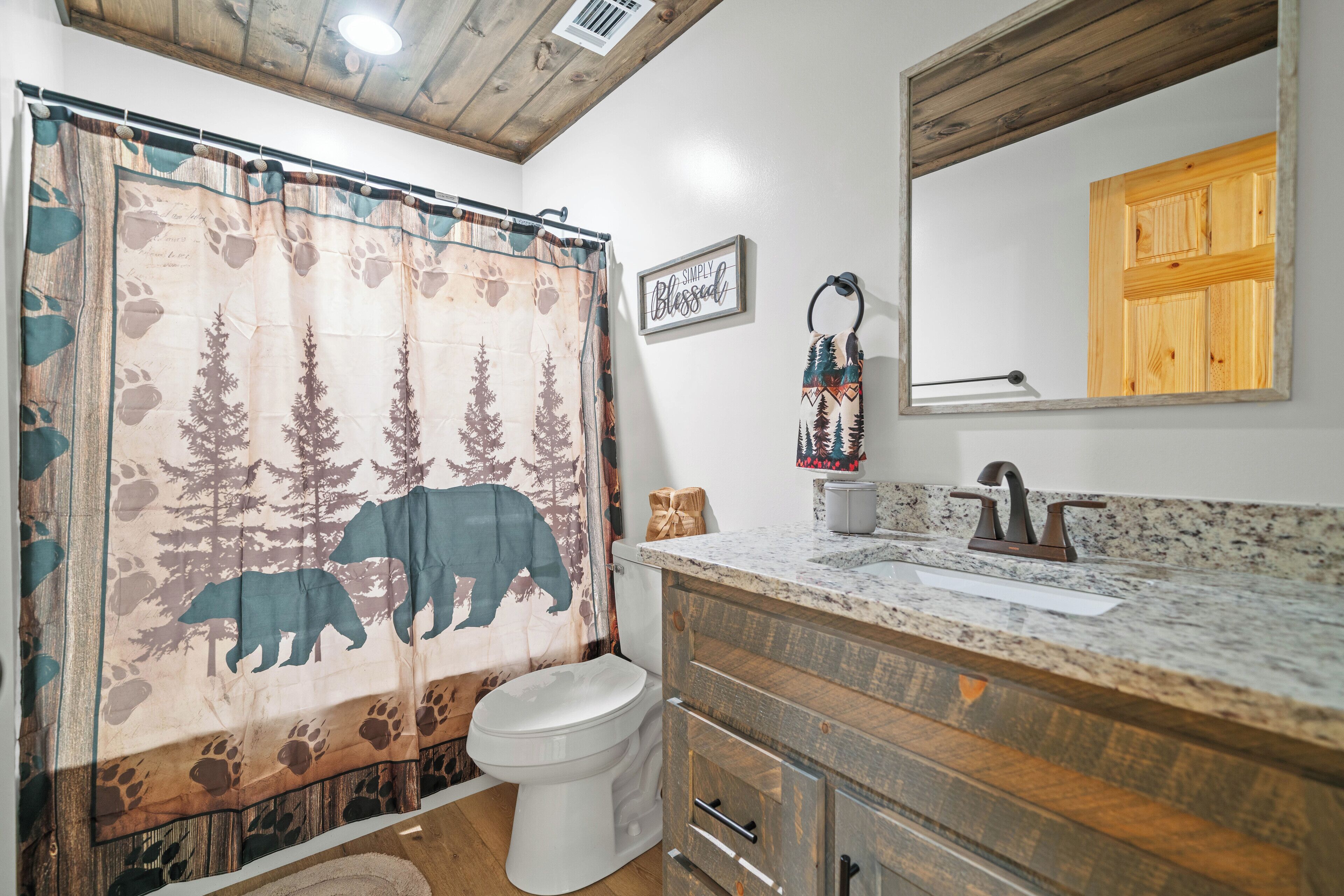 A bathroom with a wooden ceiling, a shower curtain with a forest and bear design, a toilet, a sink with a wooden vanity, and a mirror with a wooden frame.