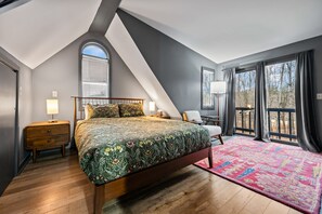 Ein Schlafzimmer mit einem großen Bett, einem Nachttisch aus Holz, einem Schreibtisch und einem Stuhl. Große Fenster mit Vorhängen bieten Blick auf Bäume.