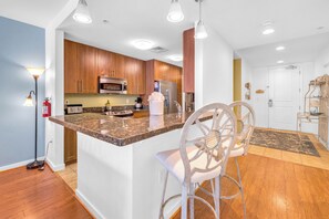 A modern kitchen with wooden cabinets, a granite countertop, and stainless steel appliances. There are two white bar stools and a fire extinguisher on the wall. - 
Sea-N-Stars is Steps to the Beach and Pool View (Virginia Beach)