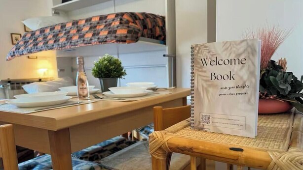 A dining area with a wooden table, chairs, a welcome book, a bottle, and a small plant. - Maison Letì Ca-Sa Check (Aprica)