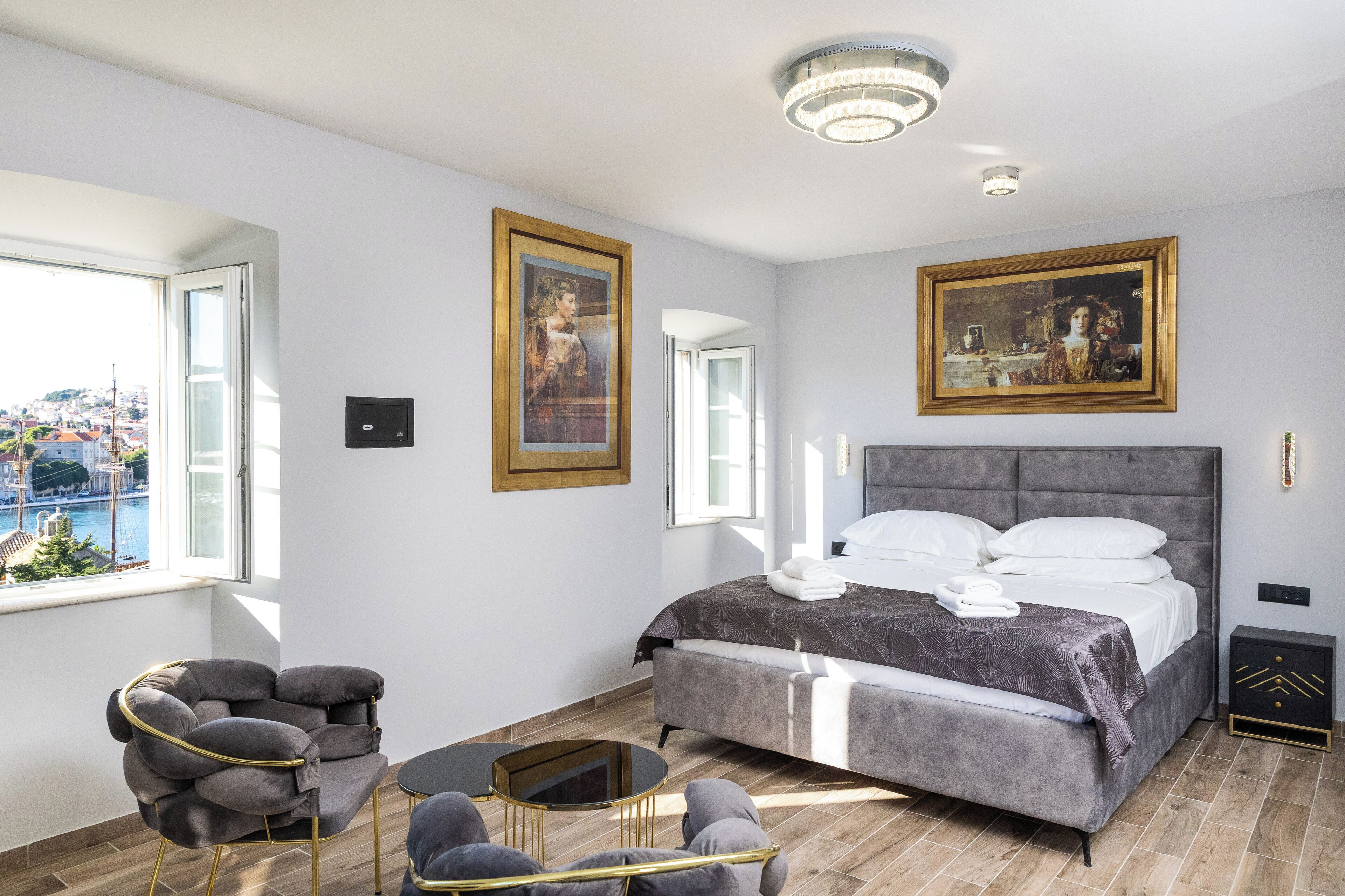 A modern bedroom with a large bed, two framed paintings, a view of a cityscape through a window, and a round coffee table.