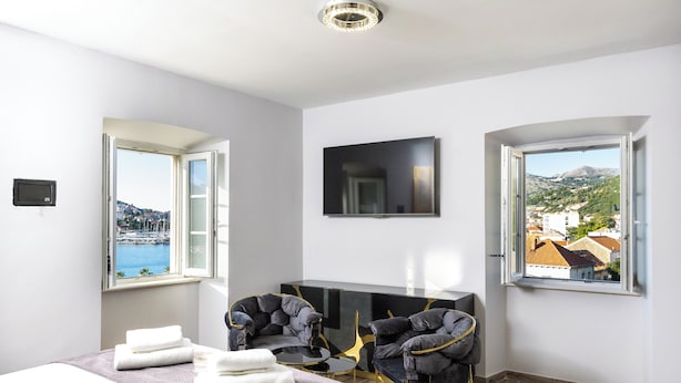 Habitación de hotel moderna con cama, dos sillones, televisor y vistas al mar y edificios.