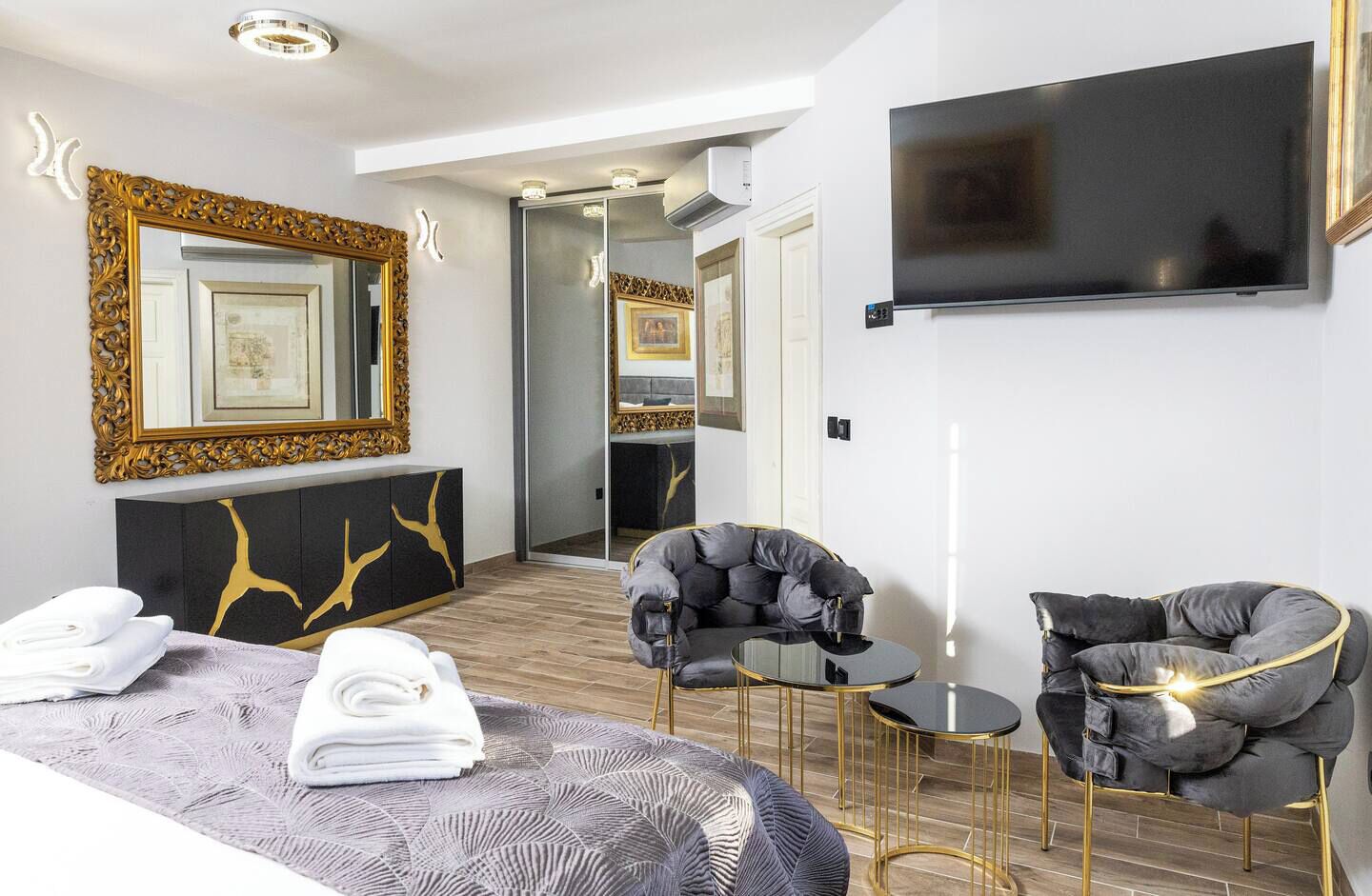 A modern hotel room with a large bed, two armchairs, a small table, a TV, and a mirror with a decorative frame.