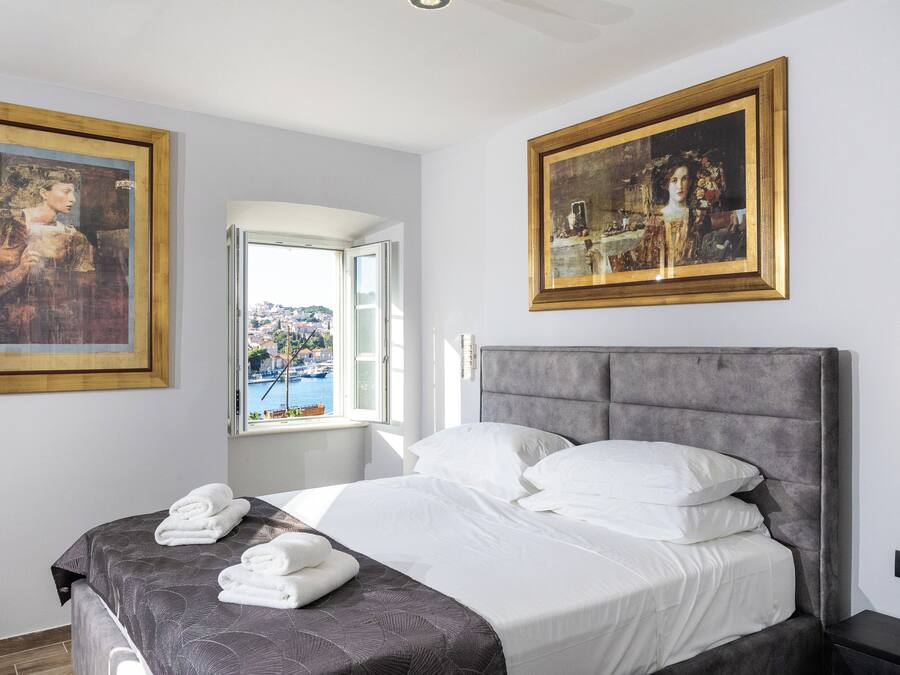 A neatly made bed with white linens and a gray headboard, a window with a view of a town, and two framed paintings on the walls.
