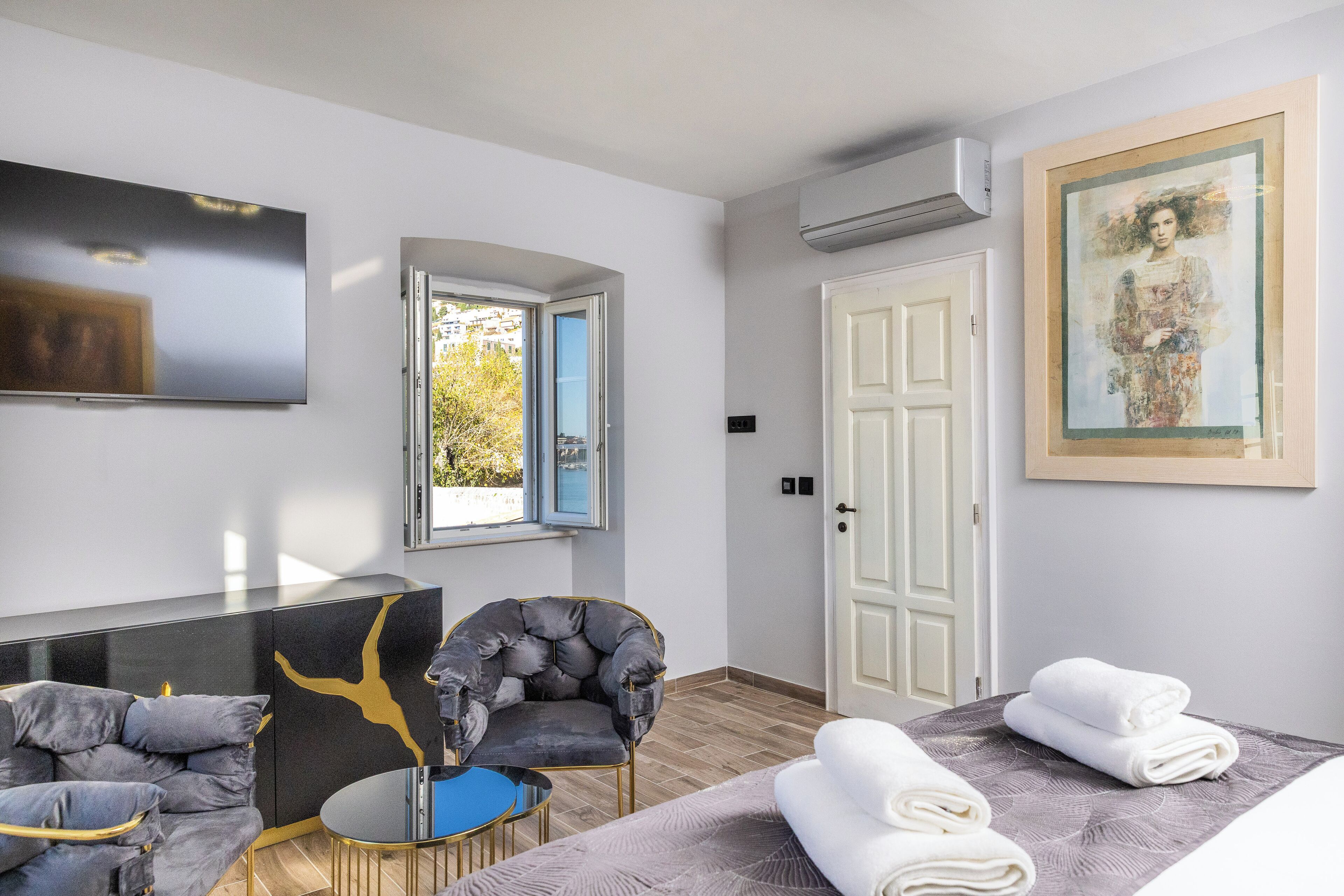 Moderni hotellihuone, jossa on litteänäytt öinen televisio, mukava nojatuoli, pieni pyöreä pöytä, siististi taitelluilla valkoisilla pyyhkeillä varustettu sänky ja suuri ikkuna, josta avautuu näkymä puita kohti.