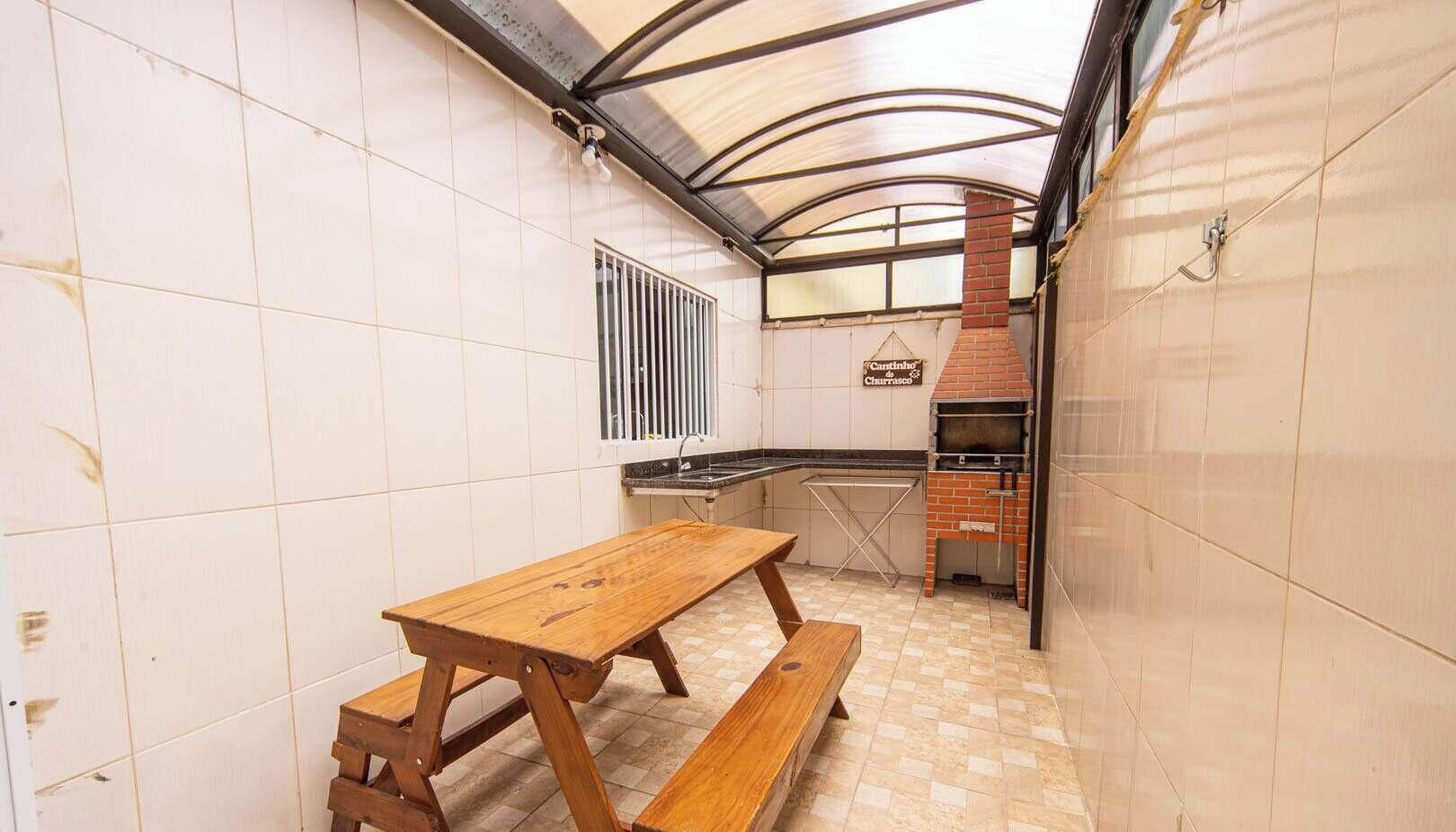 Une cuisine compacte avec une table et des bancs en bois, un four en briques et une fenêtre dotée d’une grille d’aération.