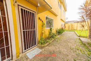 A yellow building with a barred door, a small window with a metal grill, and a paved pathway leading to a grassy area. - Casa em Mongaguá a 450m da Praia com Churrasqueira (Mongaguá)