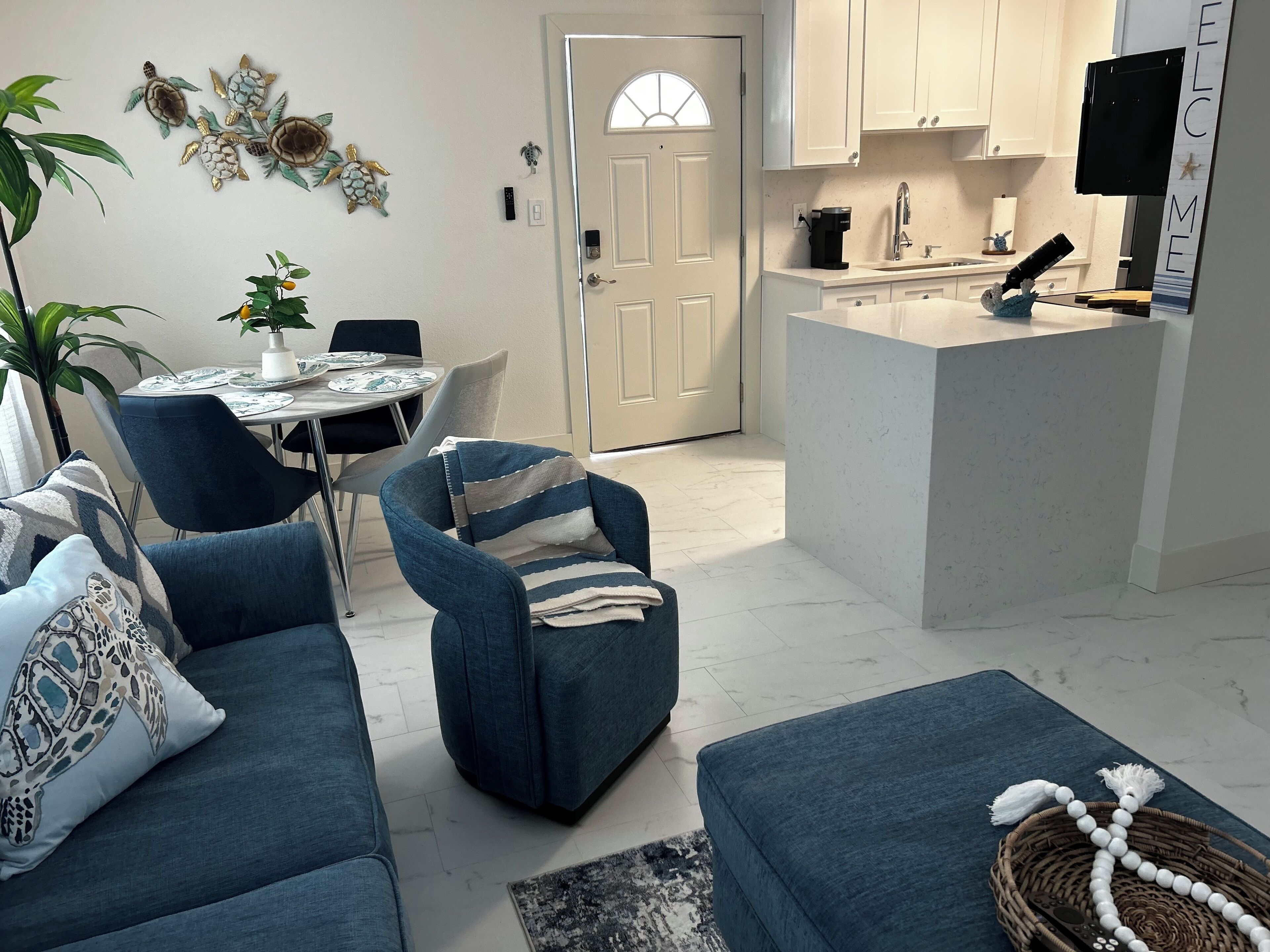 Een moderne woonkamer met een blauwe bank, een eettafel met stoelen en een keukenhoek met een koffiezetapparaat.