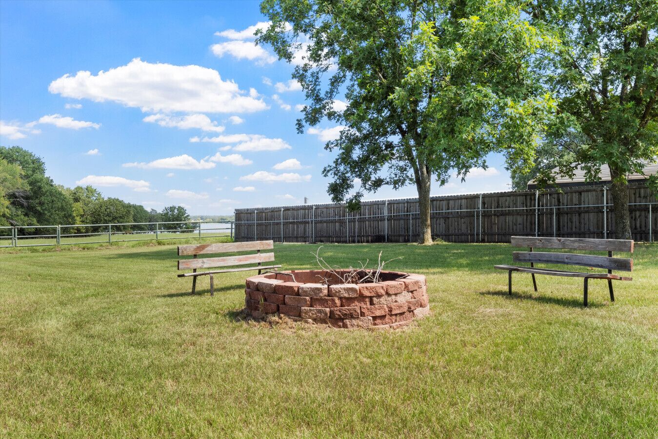 A grassy outdoor area with two wooden benches, a brick fire pit, and a wooden fence in the background.