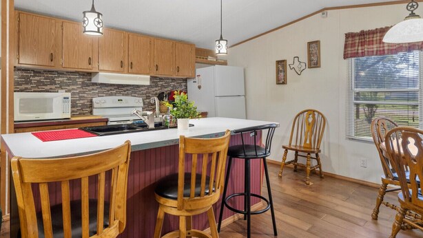 A compact kitchen with wooden cabinets, a microwave, a stove, and a dining area with wooden chairs and a table. - Lake Limestone Getaway w/ Firepit (Groesbeck)