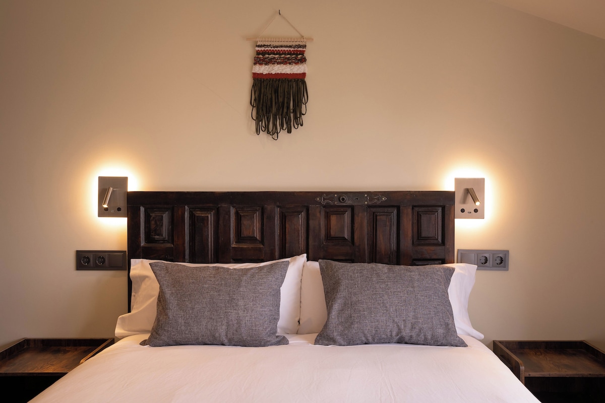 A neatly made bed with two pillows, a headboard with wall-mounted lights, and a woven wall hanging.