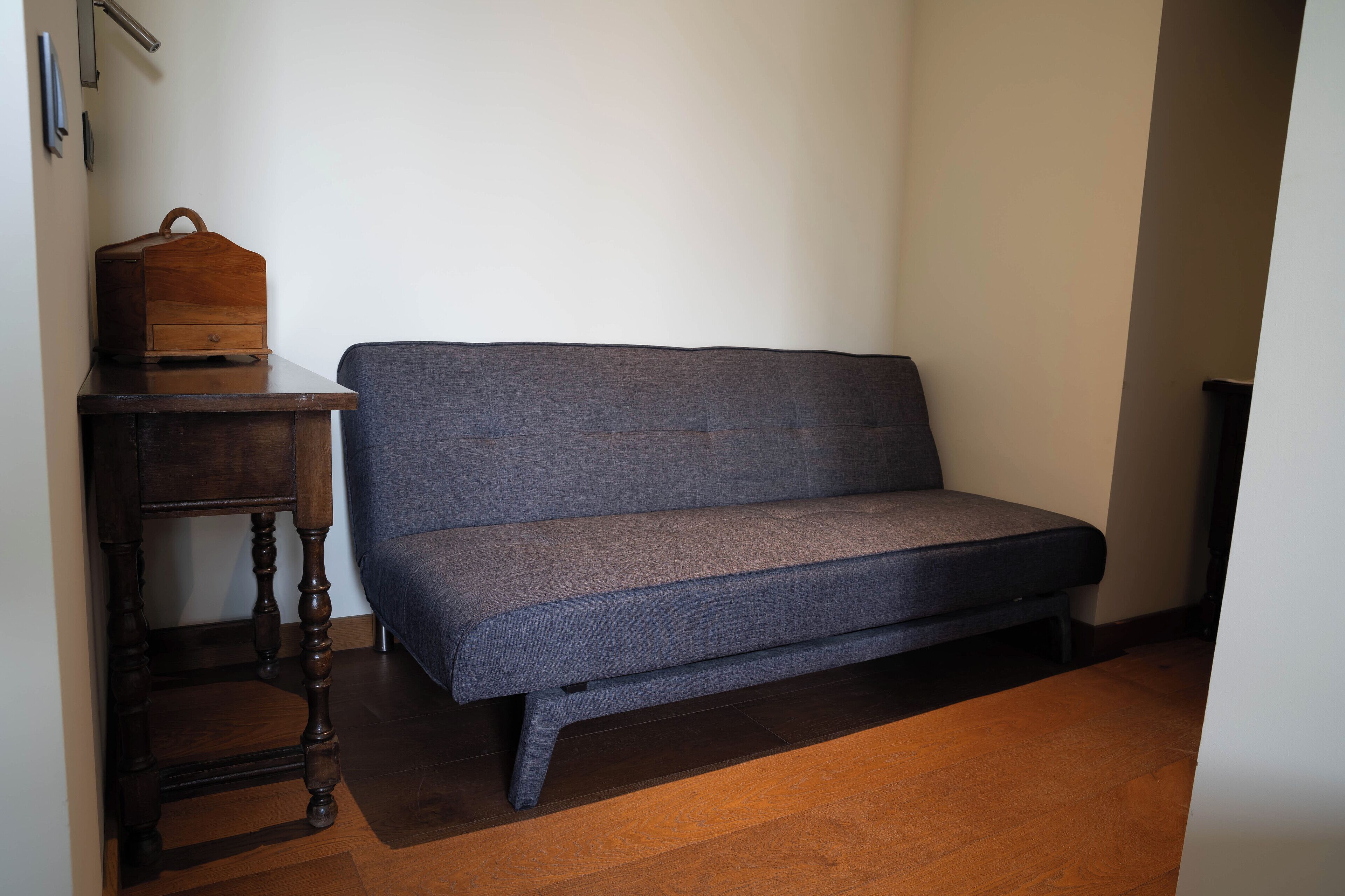 A room with a dark wooden floor, a grey sofa, a small wooden table with a drawer, and a wall-mounted shelf.