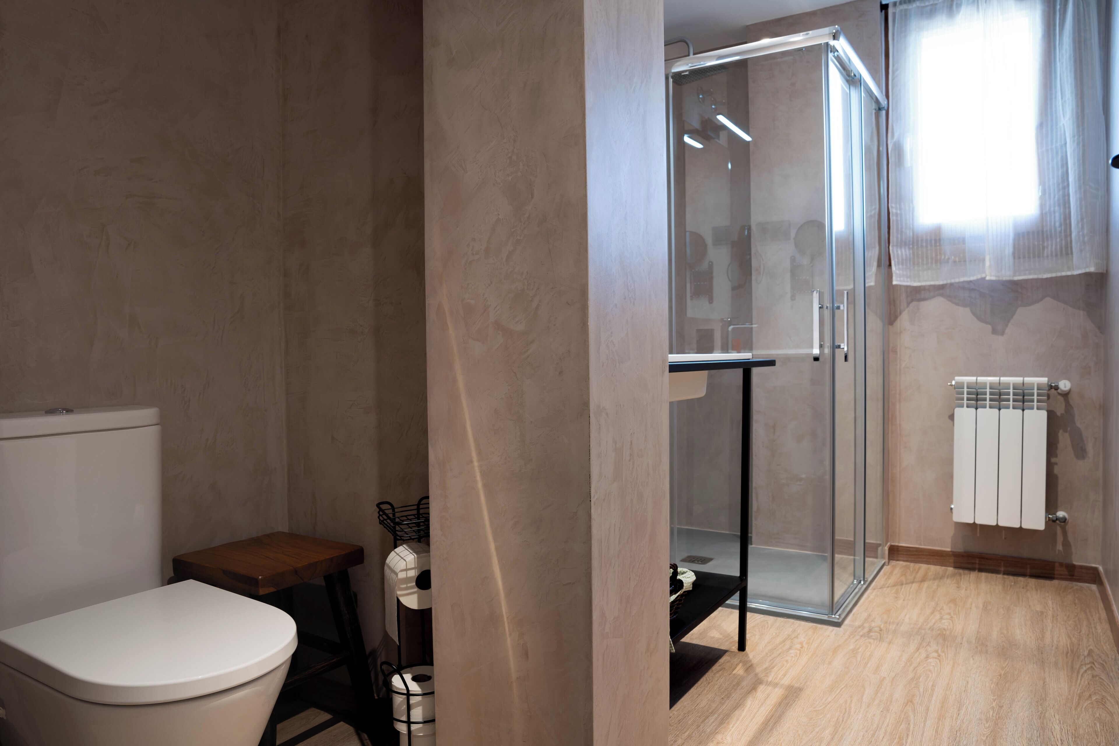A modern bathroom with a toilet, a small wooden table, a glass shower enclosure, and a radiator.