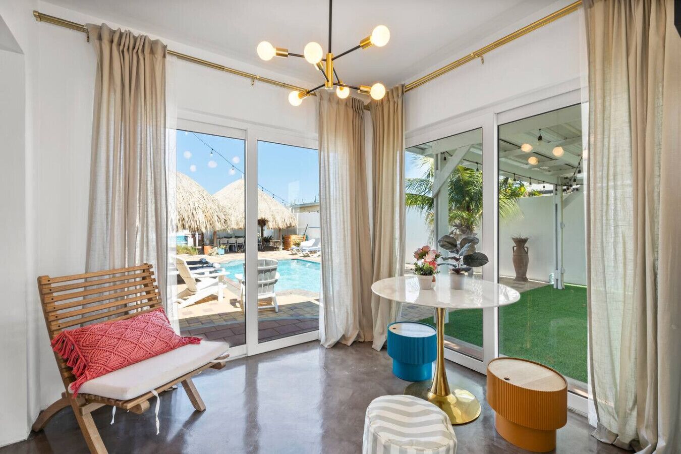 A modern room with a large glass door leading to a pool area, a round table with a blue chair, and a wooden bench with a red cushion.
