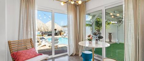 Una habitación moderna con un gran ventanal que da a una zona de piscina, una mesa redonda con una silla azul y un banco de madera con un cojín rojo.