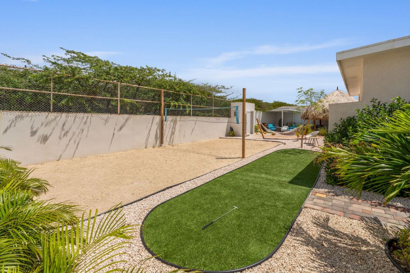 Un jardin avec un bac à sable, une pelouse artificielle, un hamac et une structure recouverte d’un toit de chaume.