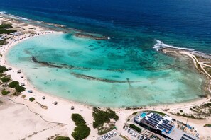 Oversigtsbillede af en strand med klart turkis vand, et stort vrag og en parkeringsplads med solpaneler.
