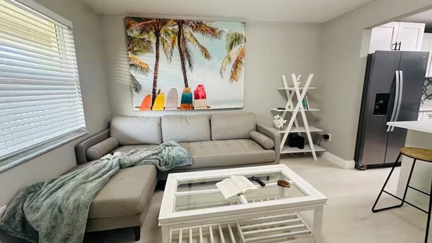 Ein Wohnzimmer mit einem beigefarbenen Sofa, einem Glastisch, einem Palmenbaum-Gemälde, einer weißen Regaleinheit und einer Küchenzeile mit einem Kühlschrank.