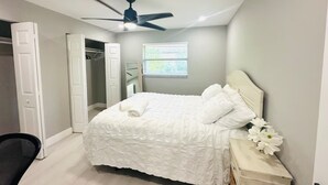 Ein Schlafzimmer mit Bett, Deckenventilator, Fenster mit Jalousien, Nachttisch mit Blumenvase und offenem Kleiderschrank.