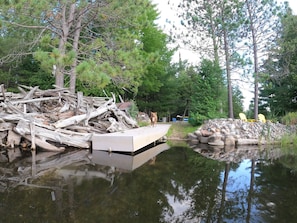 A dock extending into a calm body of water, surrounded by dense trees and a pile of logs. - Modern house on the AuSable w/ canoe & swimming access (Grayling)