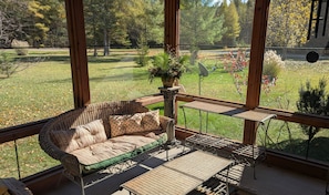 A sunlit room with a wicker sofa and a small table, large windows offering a view of a grassy yard and trees. - Modern house on the AuSable w/ canoe & swimming access (Grayling)