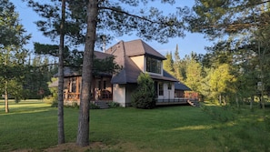 A modern two-story house with a large deck, surrounded by a well-maintained lawn and tall pine trees. - Modern house on the AuSable w/ canoe & swimming access (Grayling)