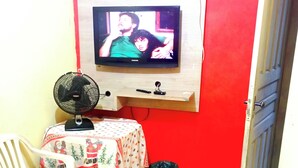 Una habitación pequeña con una televisión montada en la pared, un ventilador, una mesa con mantel temático navideño, una silla y una cama con manta roja y amarilla.