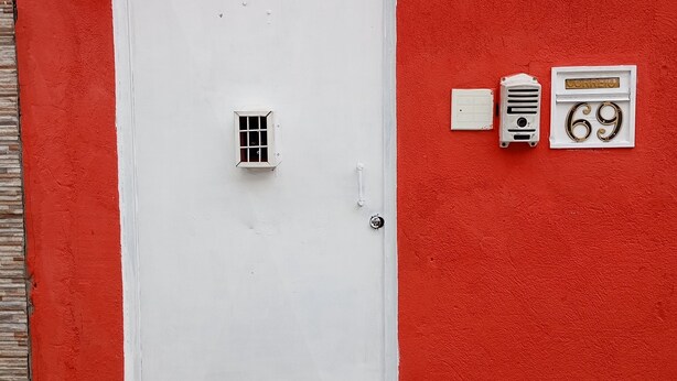 Una puerta blanca con un ventanal pequeño, un buzón y el número 69 en una pared roja.