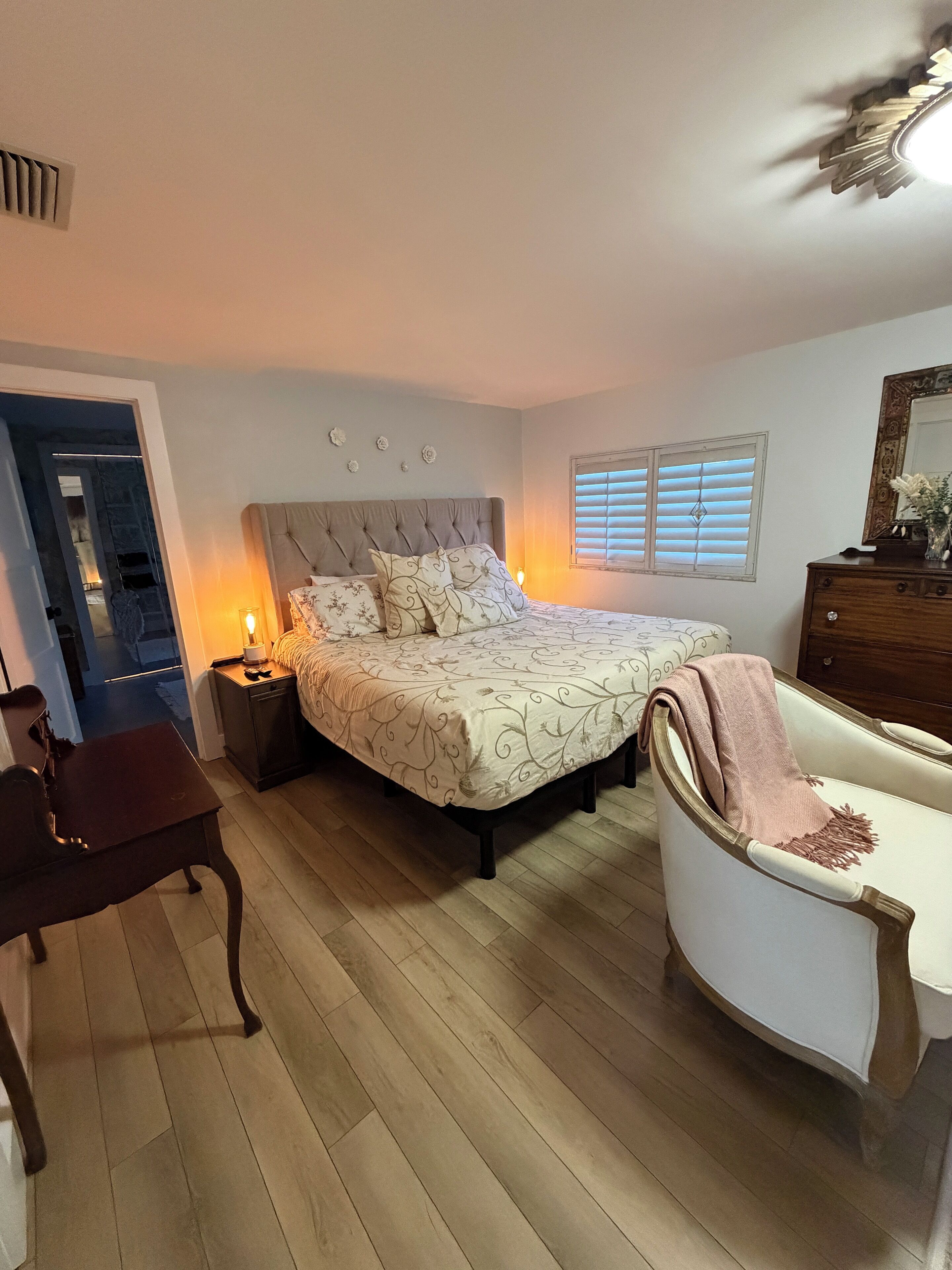 Un dormitorio con una cama, mesitas de noche, una silla, un aparador y una ventana con persianas.