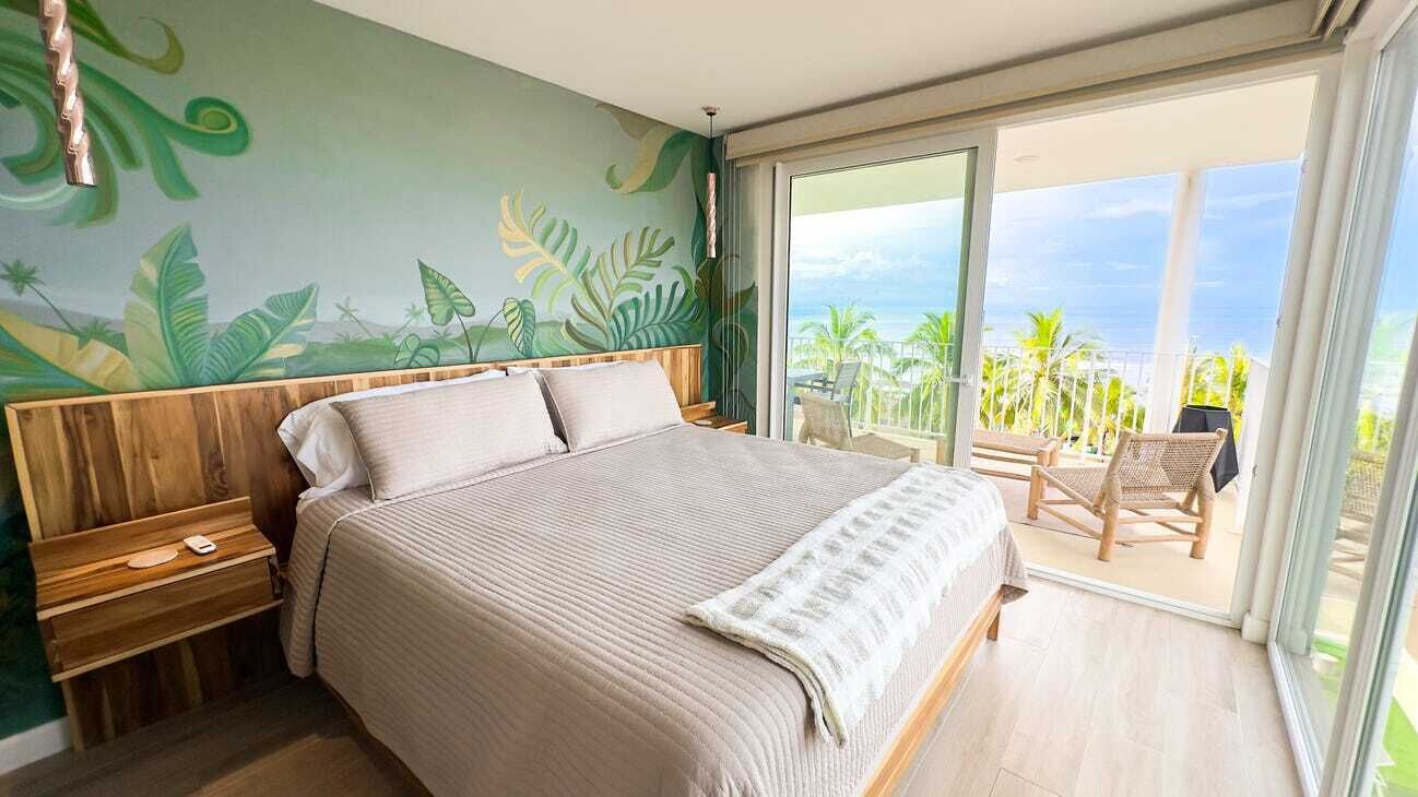 Une chambre à coucher avec un grand lit, une tête de lit en bois et une vue sur l’océan par une porte-fenêtre coulissante.