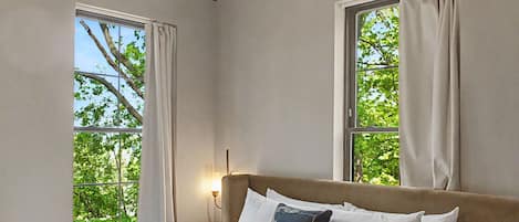 A hotel room with a large bed, two windows with curtains, a bedside table with a lamp, and a hanging light fixture.