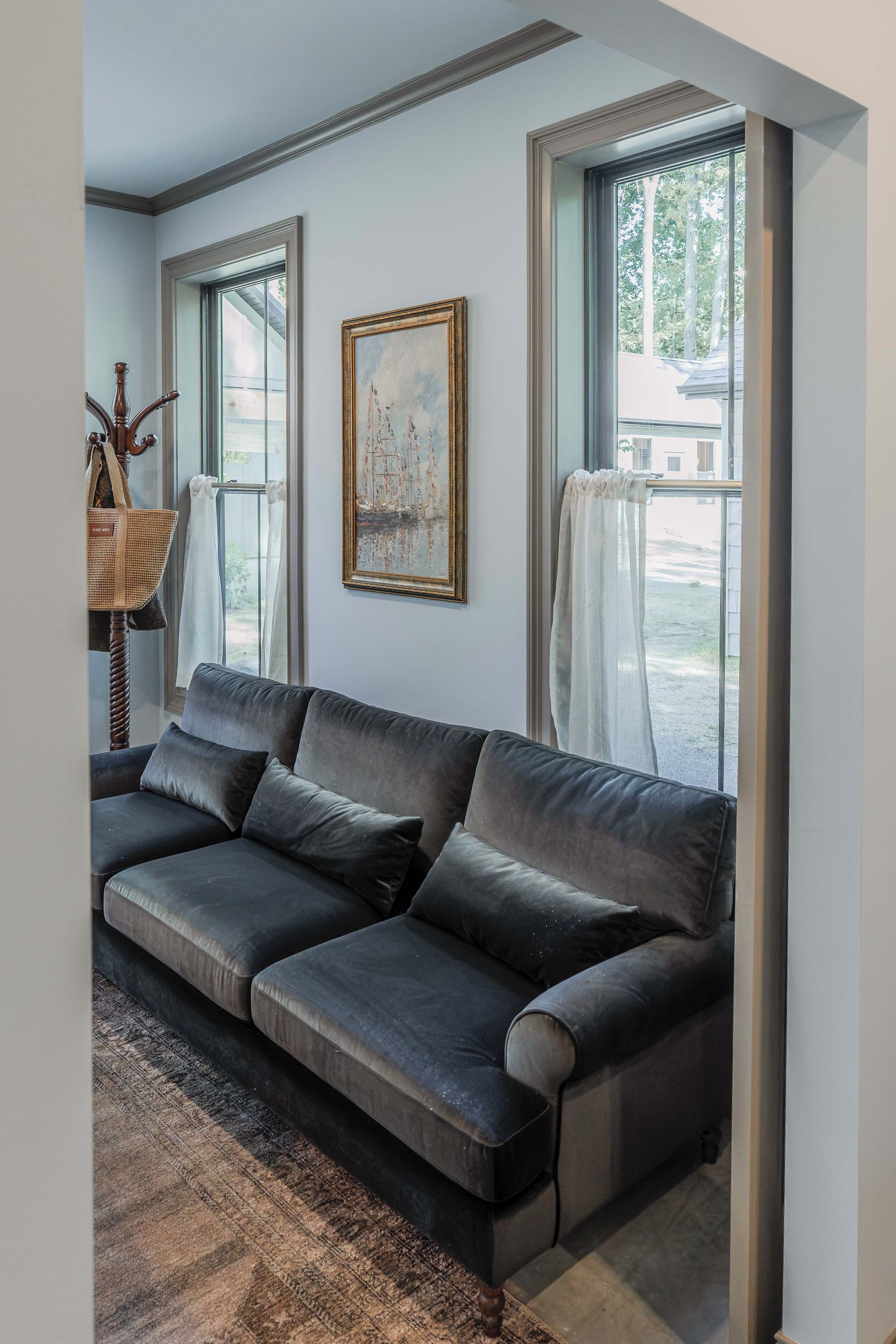 A modern living room with a dark grey sofa, a painting on the wall, and a view through large windows.