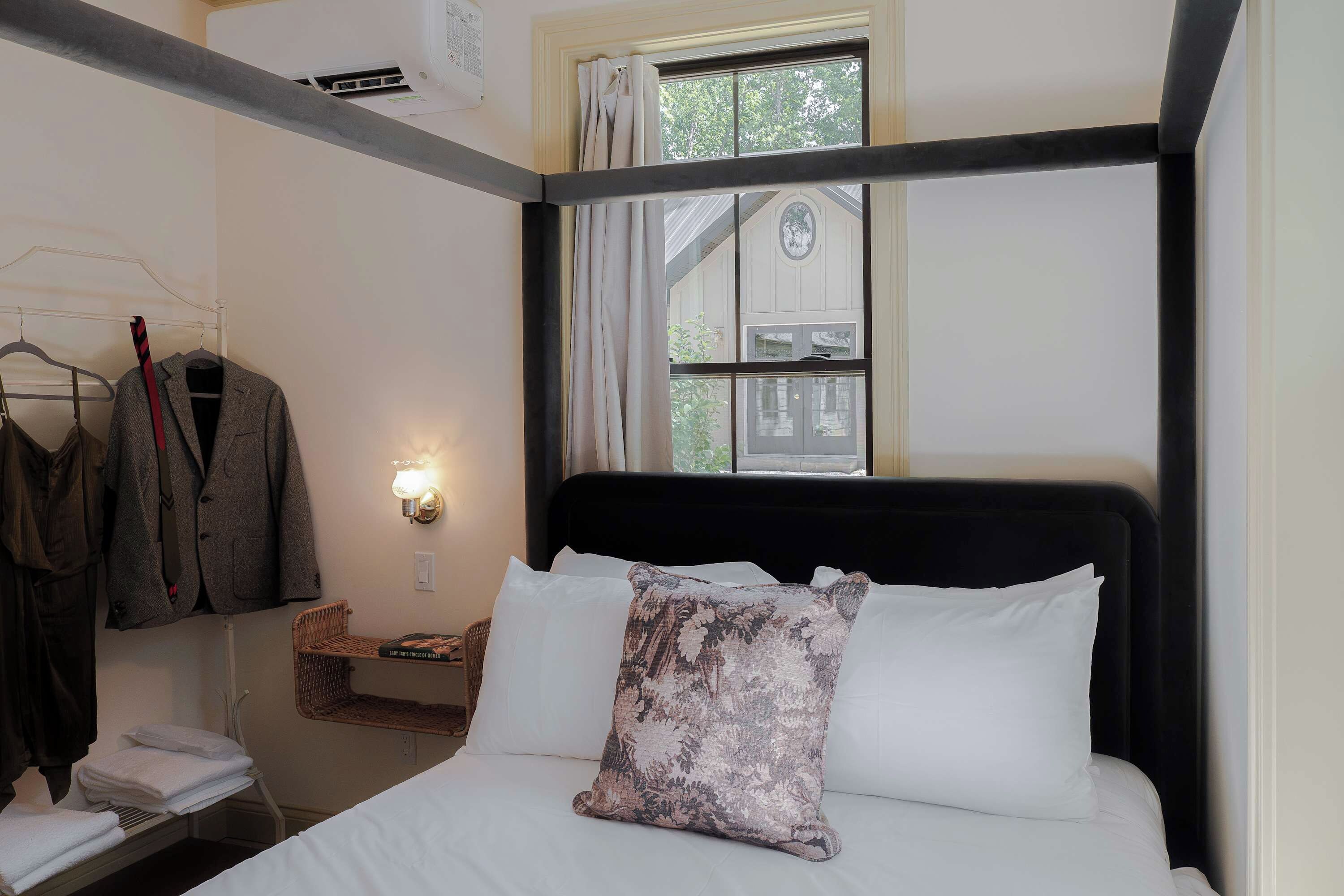 A hotel room with a bed, a nightstand, a window with curtains, a wall-mounted light, and a clothing rack.