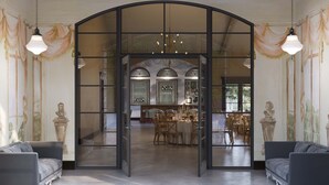 A spacious room with a high arched doorway leading to a dining area, featuring a chandelier and classical wall paintings.