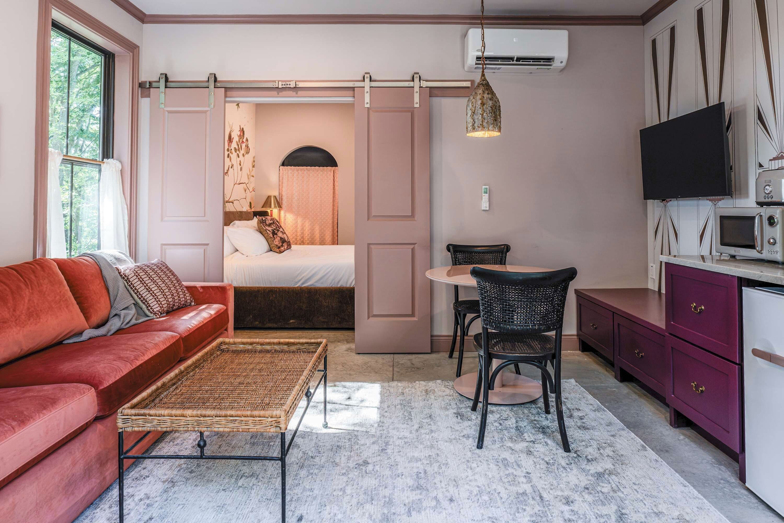 A compact room with a red sofa, a small dining table with chairs, a bed with a curtain, a hanging light fixture, a TV, and a microwave.