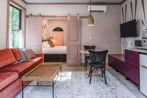 Ett litet rum med en röd soffa, ett litet matbord med stolar, en säng med ett draperi, en hängande lampa, en tv och en mikrovågsugn.