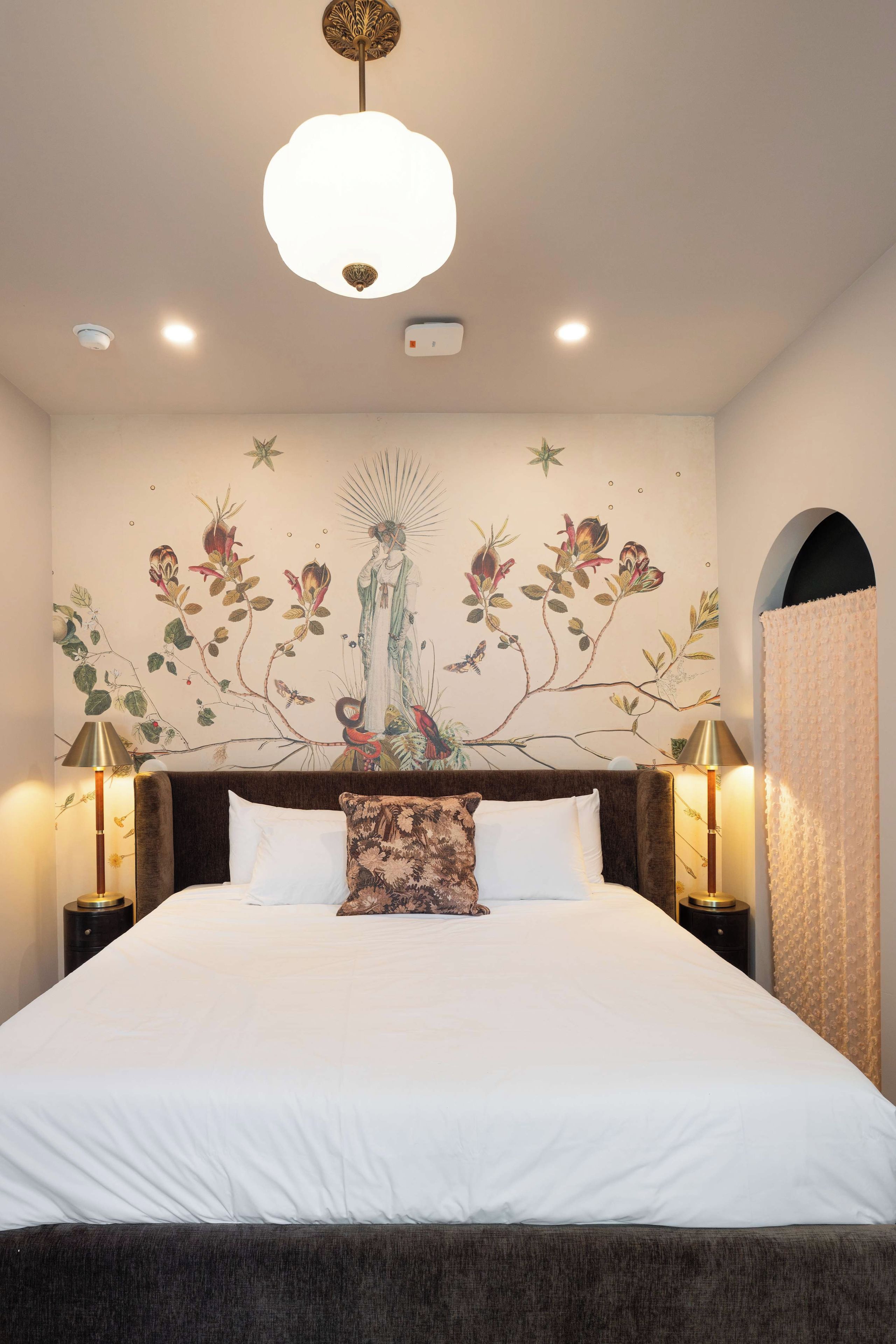 A bedroom with a large bed, two bedside lamps, a wall mural, and a ceiling light fixture.