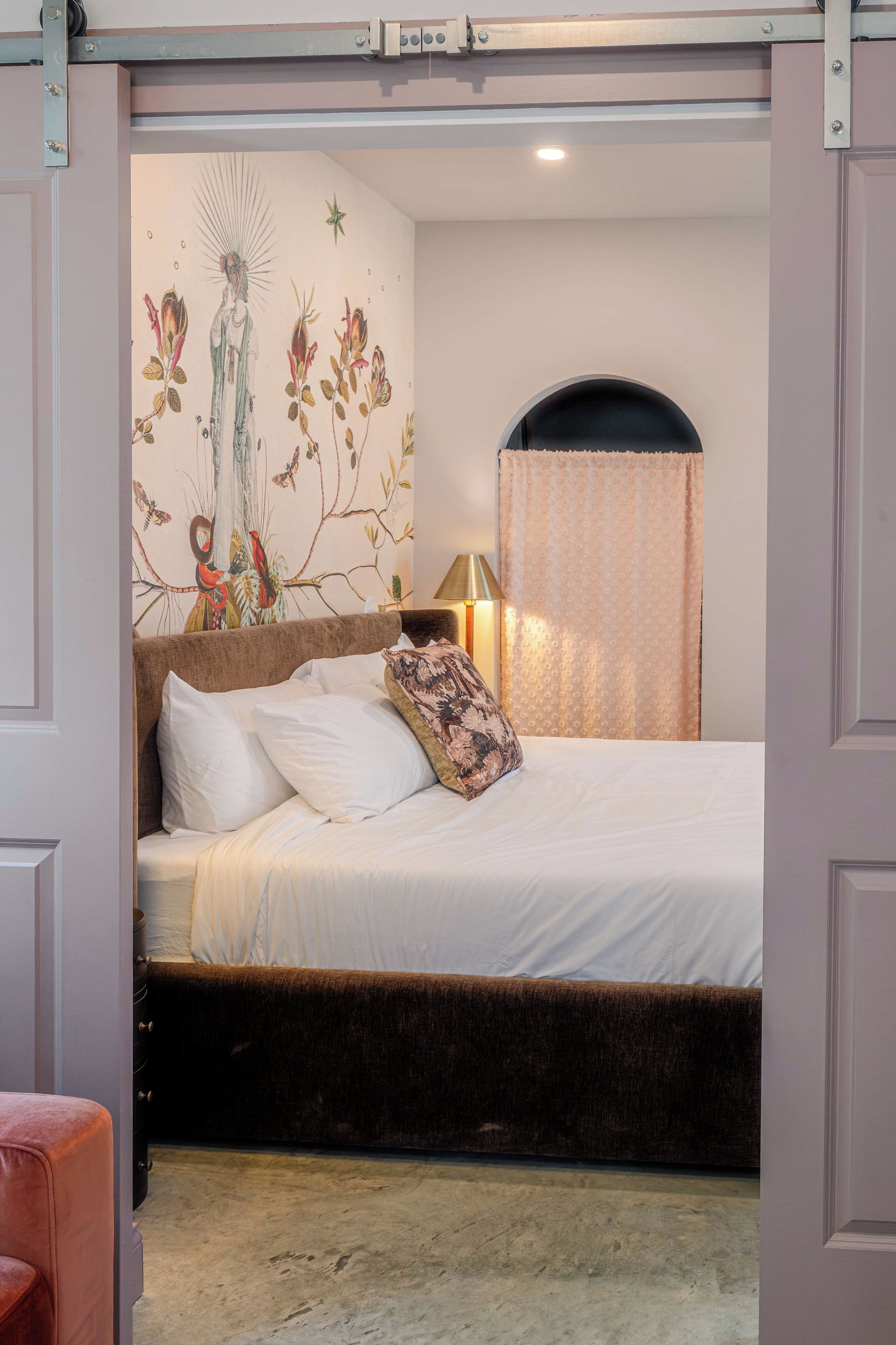 A bedroom with a bed, a nightstand, a lamp, a window with a curtain, and a wall with a floral mural.