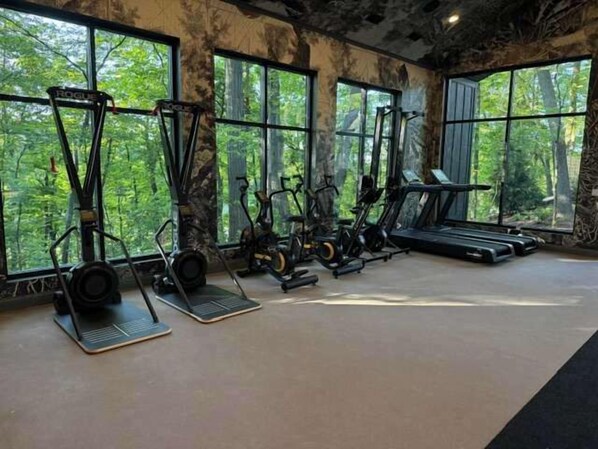 A gym with treadmills, elliptical machines, and stationary bikes, large windows with a forest view.