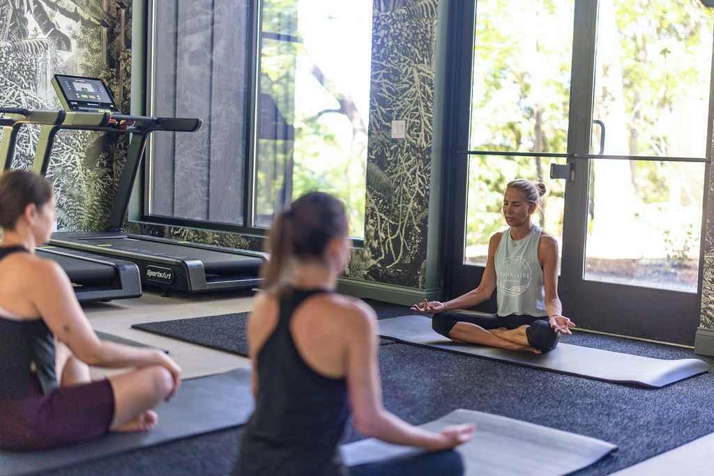 A yoga class in a gym with a treadmill and large windows.