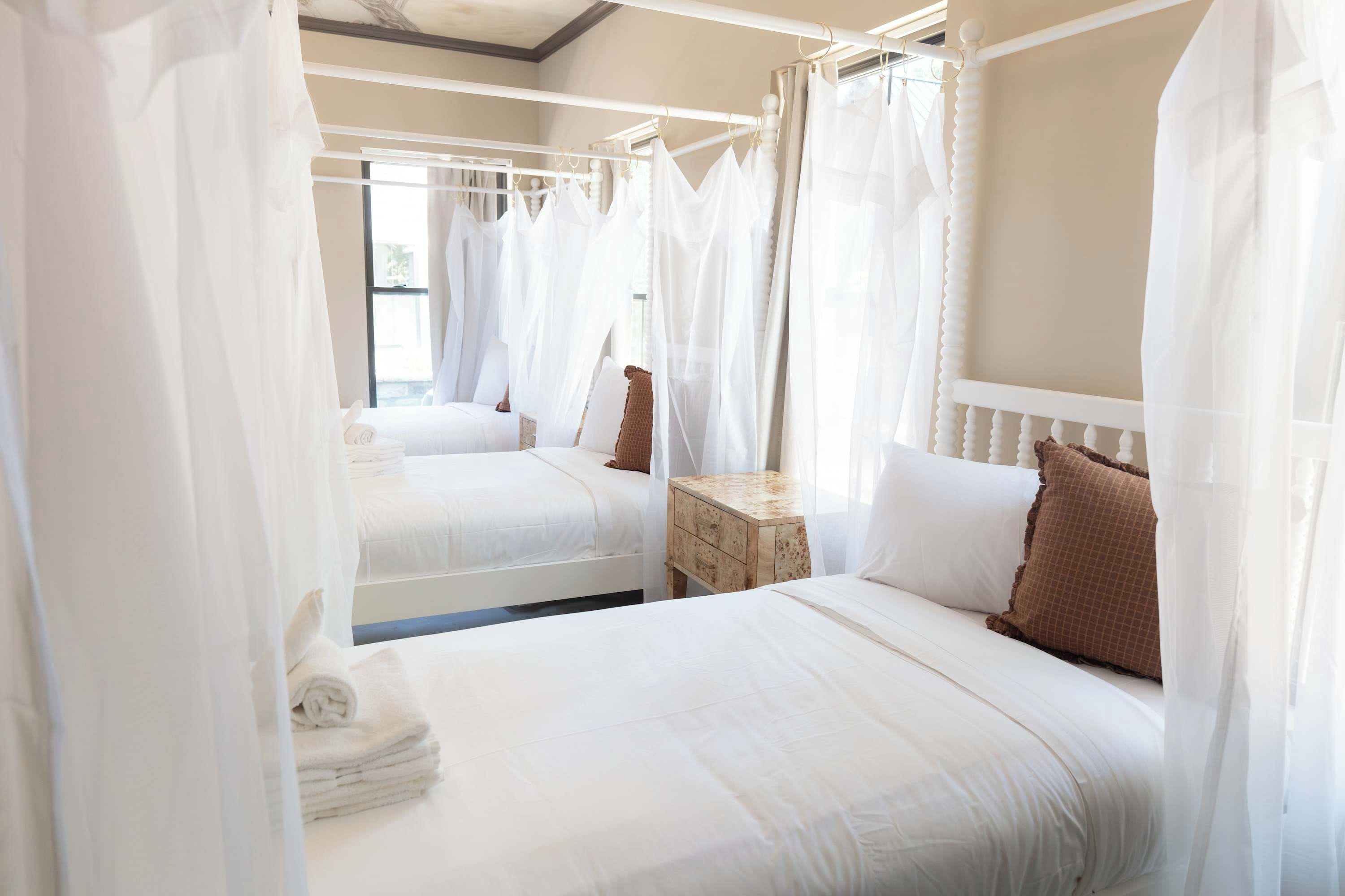 A bedroom with two beds, each with white bedding and a canopy. There is a small bedside table with a lamp.