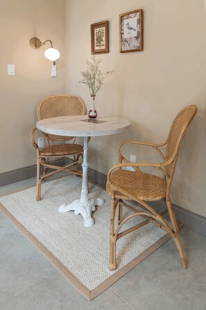 A room with a round table and two wicker chairs, a vase with dried plants, and two framed pictures on the wall.
