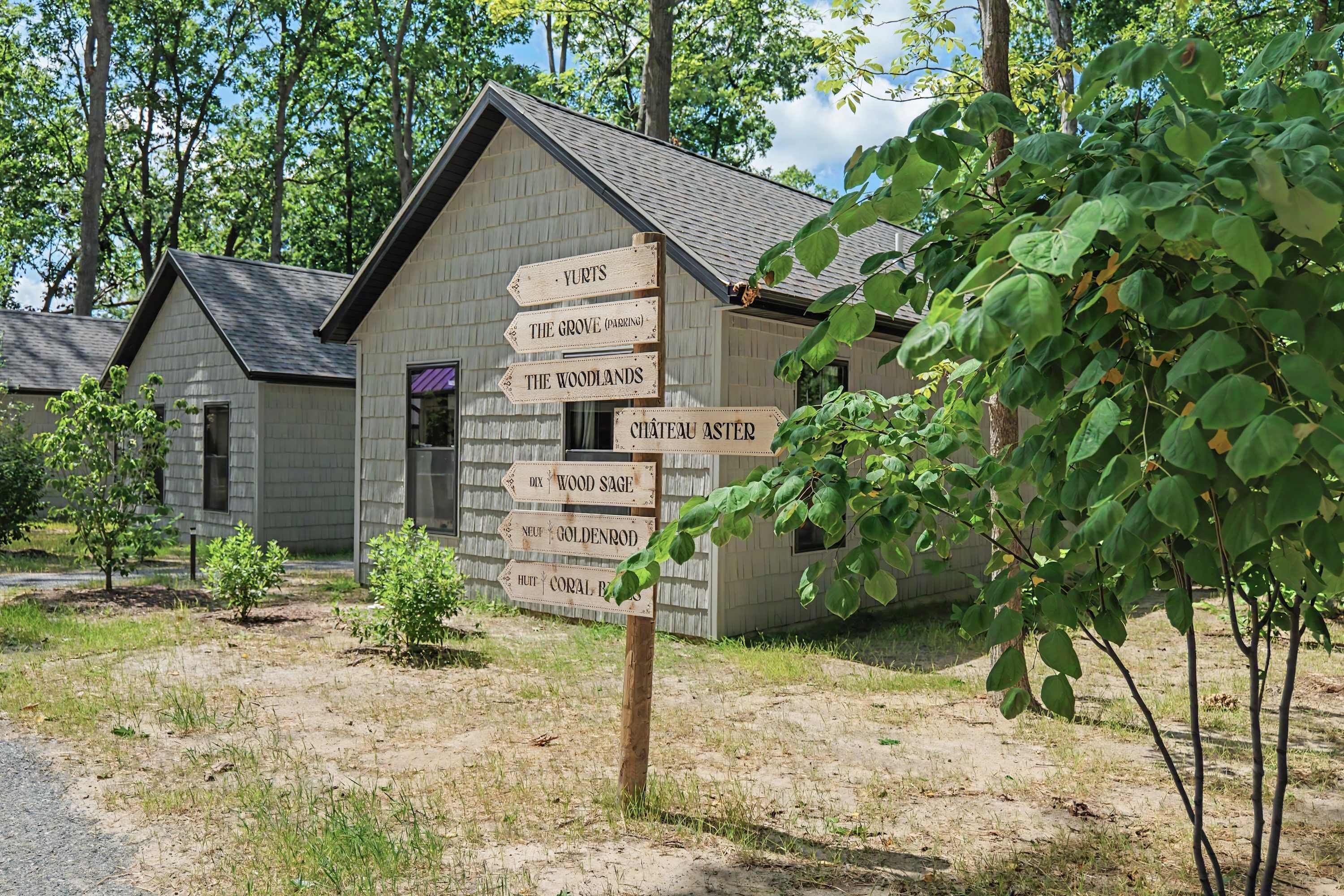 Una cabaña rústica con un cartel de madera que indica lugares cercanos como 'Yurts', 'The Grove (Parking)', 'The Woodlands', 'Chateau Aster', 'Dix Wood Sage', 'Neuf Goldenrod' y 'Huit Coral Bay'.