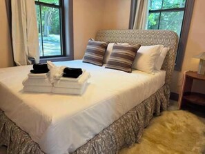 Ein ordentlich gemachtes Bett mit Kissen und gefalteten Handtüchern, ein Fenster mit Vorhängen und ein kleiner Nachttisch mit einer Lampe.