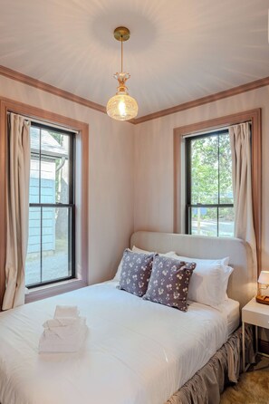 A neatly made bed with white linens and floral pillows, a bedside table with a lamp, and a view of the outdoors through two windows with curtains.