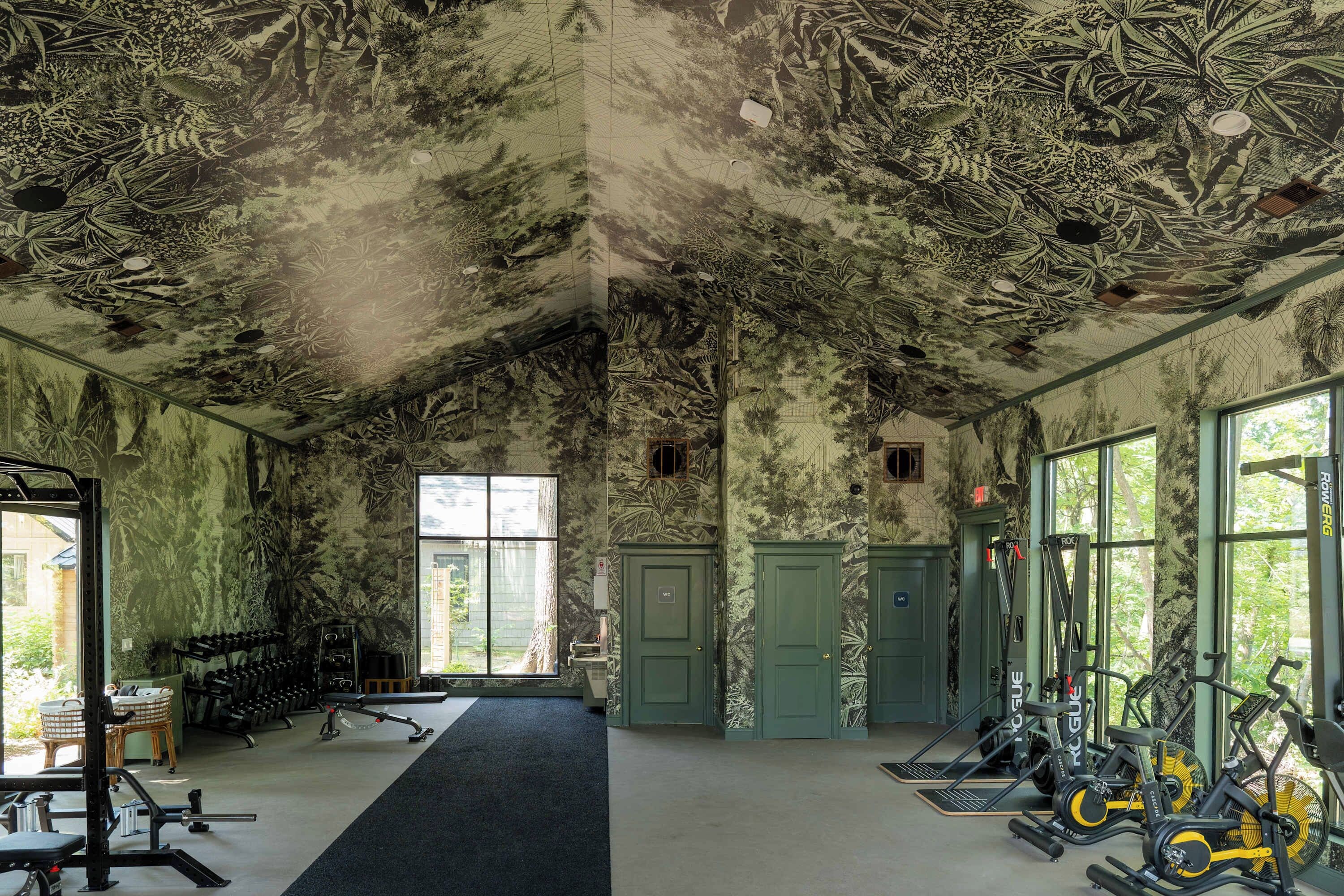 Ein Fitnessraum mit Trainingsgeräten, großen Fenstern und einer Decke mit Blätelmuster.