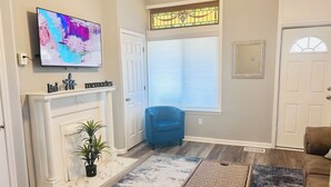 Ein Wohnzimmer mit Kamin, Fernseher, einem blauen Sessel und einem großen Fenster mit Jalousien.