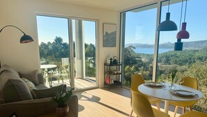 Una sala de estar moderna con un sofá, una mesa de comedor con sillas amarillas y grandes ventanales que ofrecen una vista panorámica de un bosque y un lago.